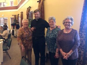 ladies in hats and gloves with Fr. Kurtis Wiedenfeld at Centennial Tea