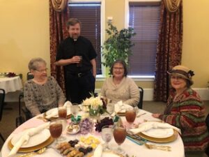ladies in hats and gloves with Fr. Kurtis Wiedenfeld at Centennial Tea